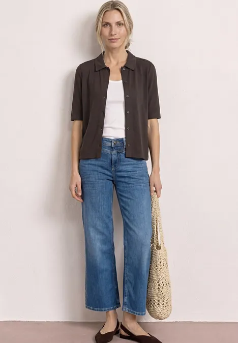 Frau trägt weiße Bluse, schwarze Kurzarm-Bluse, blaue weite Jeans und eine gehäkelte Tasche.